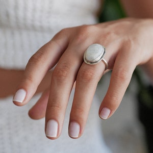 May include: A silver ring with a large oval-shaped white gemstone. The ring is set in a silver band with a textured edge.