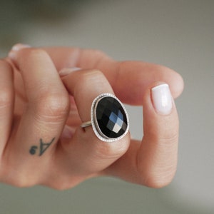 Könnte beinhalten: Ein silberner Ring mit einem schwarzen ovalen Edelstein. Der Edelstein ist facettiert und hochglanzpoliert.