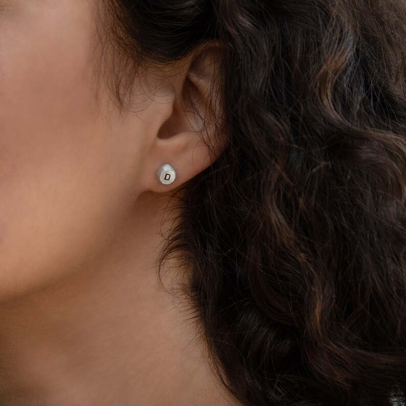 May include: A close-up of a person's ear with a small silver earring with the letter "D" on it.