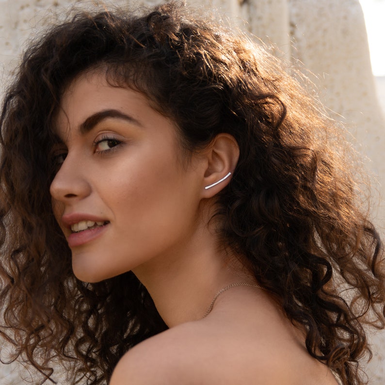 May include: A woman with curly brown hair is wearing a silver ear cuff. The ear cuff is a thin, curved bar that sits on the upper part of the ear.