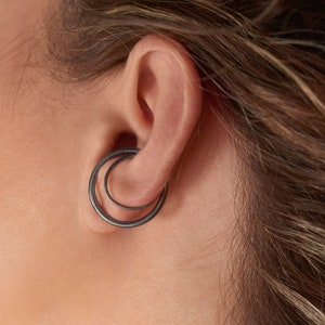 Op de afbeelding: Een paar zwarte dubbele hoepeloorbellen. De oorbellen zijn gemaakt van twee dunne metalen hoepels, waarvan er één iets groter is dan de andere, die aan elkaar zijn verbonden.