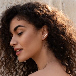 May include: A woman with long, curly brown hair is wearing a silver ear cuff on her right ear. The ear cuff is a simple, curved bar design.