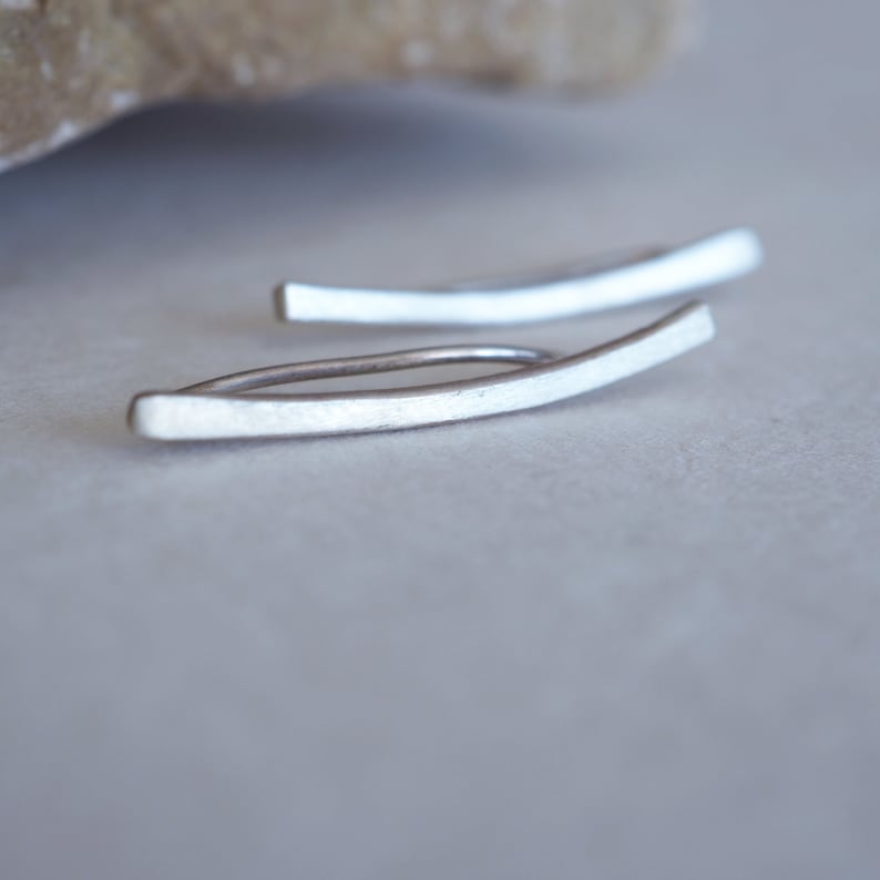 May include: A pair of silver ear cuffs, each shaped like a curved bar. The cuffs are simple and minimalist in design.