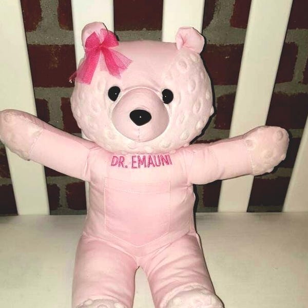 May include: A light pink plush teddy bear with a textured dimpled fabric on its head and limbs. The bear features a small pink tulle bow on its right ear, black plastic eyes, a black nose, and the text DR. EMAUNI embroidered in pink on its chest.