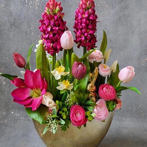 Könnte beinhalten: Eine goldene Vase gefüllt mit rosa und weißen Blumen, darunter Hyazinthen, Tulpen und Ranunkeln. Die Blumen sind symmetrisch angeordnet, mit den Hyazinthen in der Mitte und den Tulpen und Ranunkeln an den Rändern.