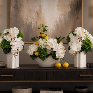May include: Three floral arrangements featuring white orchids, green hydrangeas, and yellow lemons. Two arrangements are in tall, white, vertically ribbed cylindrical vases, while the central arrangement is in a wide, white, oval-shaped bowl.