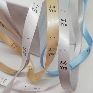 Puede incluir: Primer plano de tres cintas de satén de diferentes colores con rangos de edad impresos en ellas. Las cintas son blancas, beige claro y azul claro. El texto en las cintas dice "1-2 años", "3-4 años" y "5-6 años".