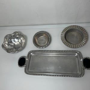 Puede incluir: Una colección de cuatro piezas de servicio de metal plateado que incluye un cuenco pequeño festoneado, un cuenco redondo con borde texturizado, un plato circular de borde ancho y una bandeja rectangular con dos detalles ovalados negros en las asas.