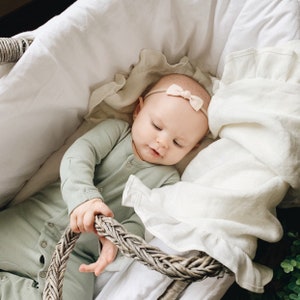 May include: A baby in a light green onesie and a beige bow headband, lying in a woven basket lined with white fabric. The baby is holding the basket's handle. The image has a soft, natural light, creating a calm and peaceful scene.