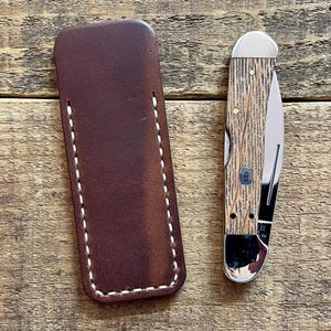 May include: A brown leather knife sheath with white stitching next to a pocket knife. The knife has a wood grain handle and a silver blade. The knife is partially open, revealing the blade. The items are on a wooden surface.