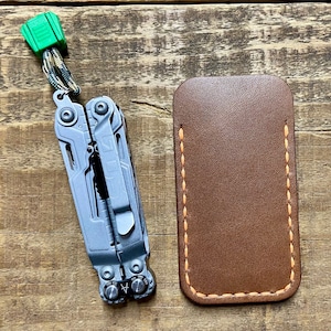 May include: A brown leather multi-tool pouch with orange stitching. The pouch is sitting on a wooden surface next to a silver multi-tool with a green lanyard.