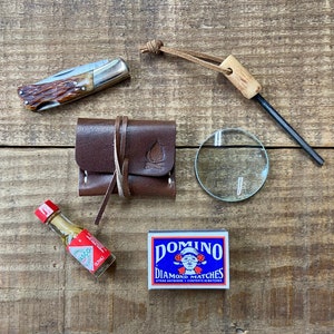 May include: A collection of outdoor survival gear on a wooden surface. Includes a pocket knife, leather pouch with a campfire logo, magnifying glass, fire starter, small bottle of hot sauce, and a box of Domino Diamond Matches.