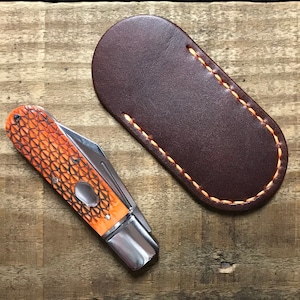 May include: A small pocket knife with an orange and black patterned handle and a silver blade. The knife is lying on a wooden surface next to a brown leather sheath with orange stitching.