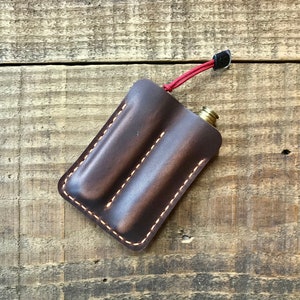 May include: A dark brown leather flask case with a red lanyard. The case has two compartments and is stitched with light-coloured thread. The flask's brass top is visible. The case is on a wooden surface.