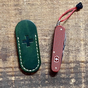 May include: A red Swiss Army knife with a white cross emblem and a green leather sheath with a cross design. The knife has a red lanyard and a black end piece. The items are on a wooden surface.