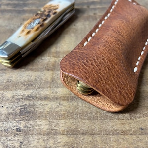 May include: A pocket knife with a cream-colored handle and a brown leather sheath. The sheath is stitched with white thread and holds several brass coins. The knife is partially open.