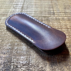 May include: A dark brown leather pen sleeve with a smooth, rounded shape and visible white stitching along the edges. The leather has a slight sheen and natural grain texture, featuring a curved flap design that secures the contents inside.