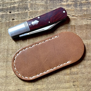 May include: A pocket knife with a dark wood handle and silver accents, resting above a brown leather sheath. The knife features a silver blade and decorative inlays. The sheath is oval-shaped with white stitching.