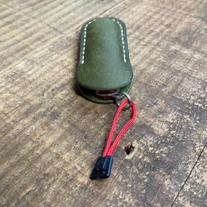 May include: An olive green leather multi-tool case with white stitching. The case is attached to a red lanyard with a black clasp. The multi-tool is visible inside the case.