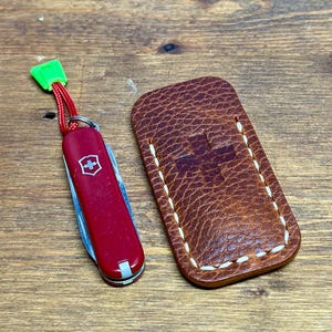 May include: A red multi-tool pocket knife with a silver cross emblem and a bright green plastic pull tab on a red cord, positioned next to a brown textured leather pouch featuring a stitched cross design and white thread detailing.