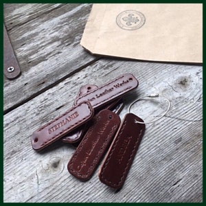 May include: A close-up of several brown leather keychains with the text "Jun Leather Works" and "Cabin Leather Works" on them. The keychains are on a wooden surface.