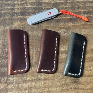 May include: A silver pocket knife with a red cross emblem and an orange lanyard is above three leather sheaths. The sheaths are brown, dark brown, and black, each with white stitching. The items are on a wooden surface.