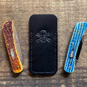 May include: Two pocket knives with folding blades and textured handles, one brown and yellow, the other blue. A black leather sheath with a skull and crossbones design is centered between the knives. The knives and sheath are on a wooden surface.