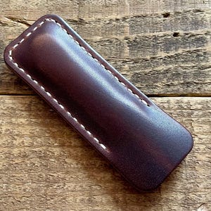 Peut inclure: Un étui en cuir bordeaux foncé avec des coutures blanches. L'étui rectangulaire a des bords arrondis et est probablement conçu pour contenir un stylo ou un autre petit objet. Le cuir a une finition lisse et polie.