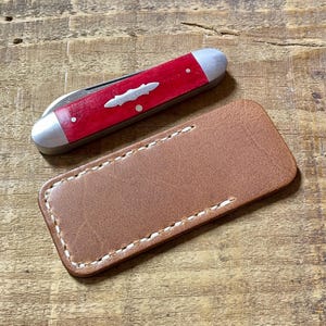 May include: A red pocket knife with a silver blade and accents, next to a brown leather sheath. The knife has a decorative silver emblem and white dots. The sheath is rectangular with white stitching.