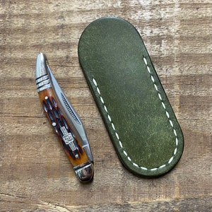 May include: A pocket knife with a brown handle and a silver blade, next to a green leather sheath with white stitching. The knife has a decorative emblem on the handle.