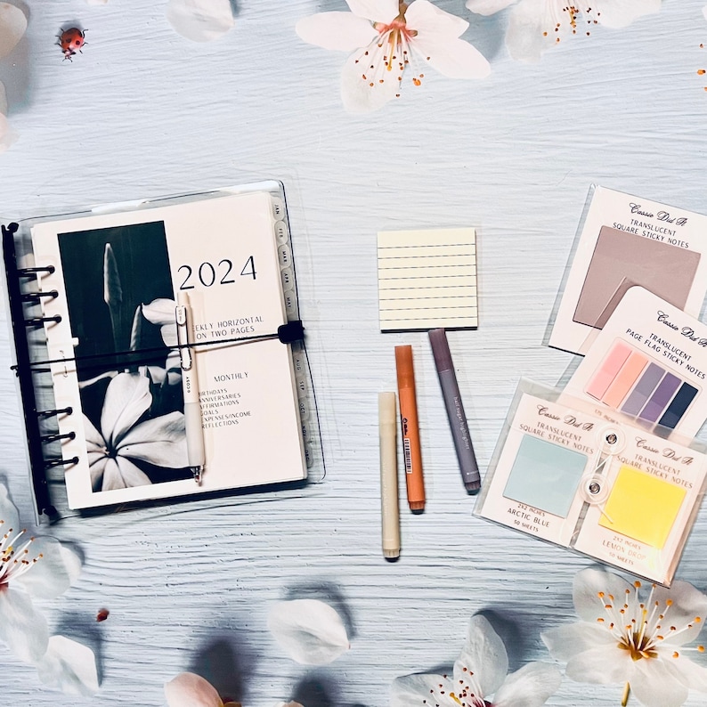 May include: A 2024 planner with a floral design on the cover, a stack of sticky notes in various colors and sizes, and a few highlighters. The sticky notes are labeled "Translucent Square Sticky Notes" and "Translucent Page Flag Sticky Notes".