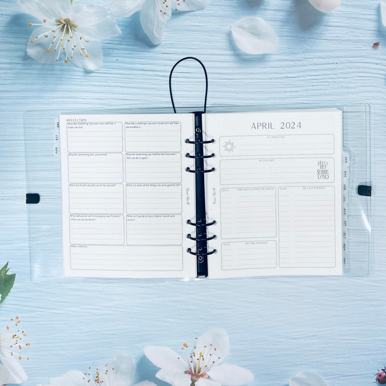 May include: A white planner with a clear plastic cover. The planner is open to the April 2024 calendar page. The calendar page has a section for appointments, reminders, and notes. The planner also has a reflection section with prompts for self-reflection.