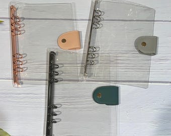 May include: Clear plastic binders with metal ring mechanisms. The binders have colored closures in peach, gray, and teal. The ring mechanisms are in rose gold, silver, and black.