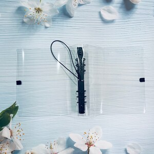 May include: A clear plastic binder with a black metal ring mechanism and a black elastic closure. The binder is lying on a light blue wooden surface.