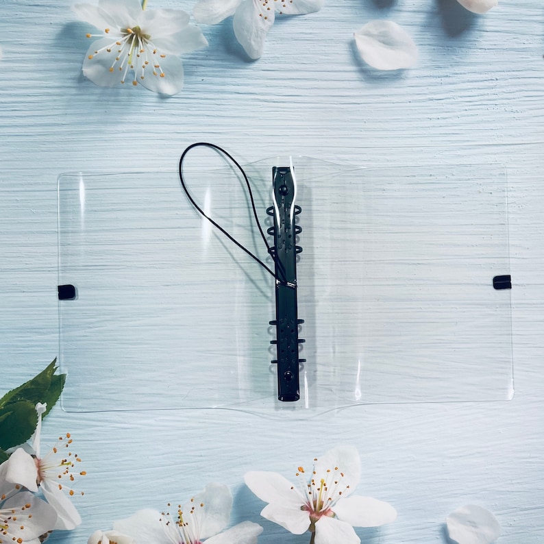 May include: A clear plastic binder with a black metal ring mechanism and a black elastic closure. The binder is lying on a light blue wooden surface.