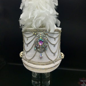 May include: A white feather plume sits atop a white hat with a silver trim and a large iridescent jewel in the center. The hat is decorated with silver chains and beads.