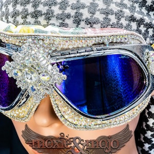 Puede incluir: Gafas estilo aviador con lentes tintadas en azul, montura plateada y un adorno central de cristal. Los bordes est&aacute;n incrustados con peque&ntilde;as perlas y pedrer&iacute;a transparente, complementadas con una cinta el&aacute;stica de color dorado.