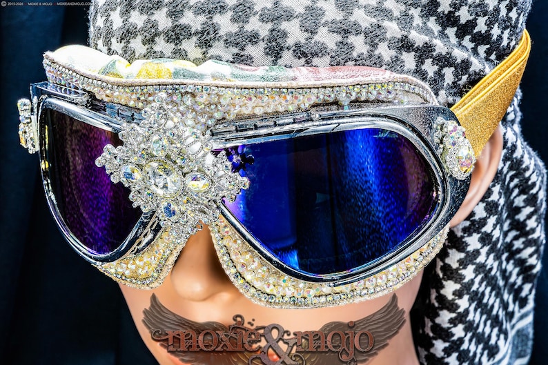 Puede incluir: Gafas estilo aviador con lentes tintadas en azul, montura plateada y un adorno central de cristal. Los bordes est&aacute;n incrustados con peque&ntilde;as perlas y pedrer&iacute;a transparente, complementadas con una cinta el&aacute;stica de color dorado.