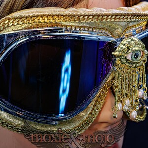 May include: Goggles with dark reflective lenses, a gold-textured frame, and a gold chain trim. A gold ornament shaped like an eye with dangling gold chains and small white pearls is attached to the side. The text moxie & mojo is visible below.