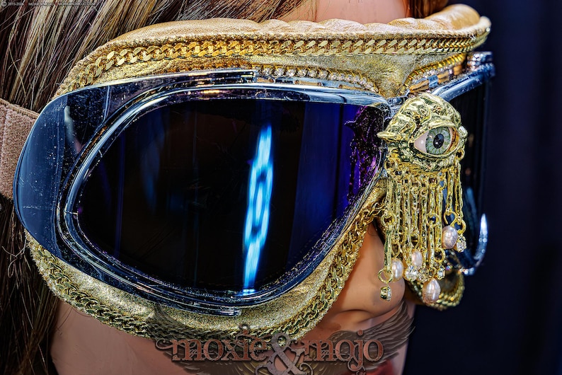 May include: Goggles with dark reflective lenses, a gold-textured frame, and a gold chain trim. A gold ornament shaped like an eye with dangling gold chains and small white pearls is attached to the side. The text moxie & mojo is visible below.