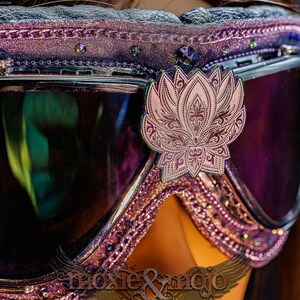 Puede incluir: Gafas protectoras de color morado con un emblema de flor de loto met&aacute;lico centrado en el puente. El marco est&aacute; decorado con purpurina morada, peque&ntilde;os cristales y un borde texturizado. Las lentes son oscuras, reflectantes y tintadas.