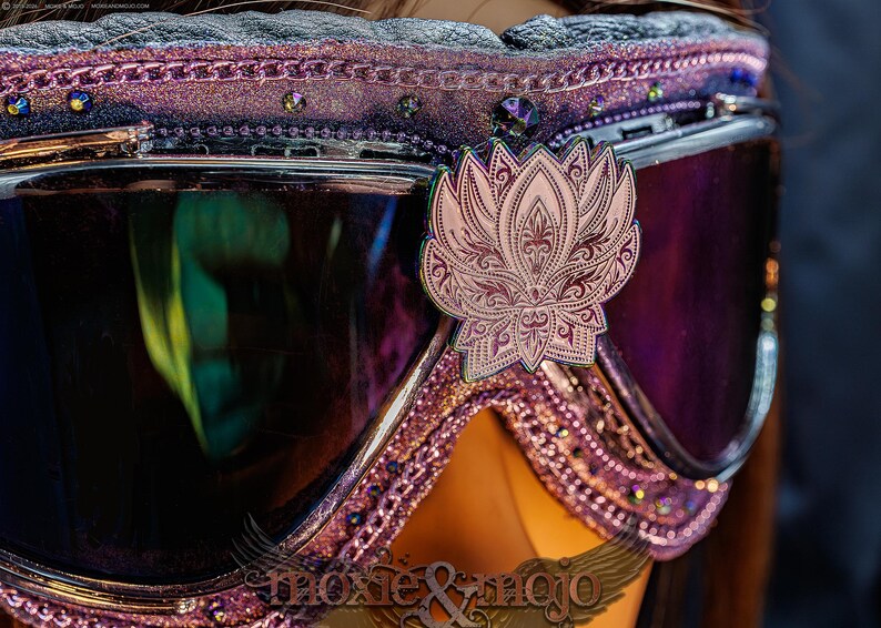 Puede incluir: Gafas protectoras de color morado con un emblema de flor de loto met&aacute;lico centrado en el puente. El marco est&aacute; decorado con purpurina morada, peque&ntilde;os cristales y un borde texturizado. Las lentes son oscuras, reflectantes y tintadas.