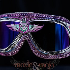 Puede incluir: Gafas protectoras con lentes reflectantes de color morado oscuro y marco plateado. El borde est&aacute; decorado con una cadena morada. Un emblema central muestra un oso con sombrero de copa y alas extendidas a los lados en tonos morado y verde azulado.