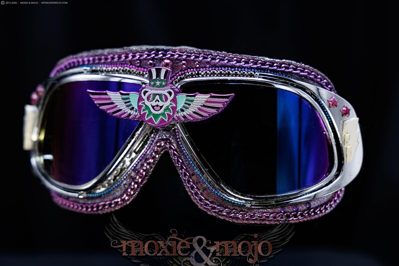 Puede incluir: Gafas protectoras con lentes reflectantes de color morado oscuro y marco plateado. El borde est&aacute; decorado con una cadena morada. Un emblema central muestra un oso con sombrero de copa y alas extendidas a los lados en tonos morado y verde azulado.