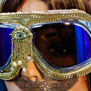 May include: Goggles with dark purple lenses, a gold-toned frame, and a decorative gold eye emblem featuring dangling white pearls and clear crystals. The frame is accented with a gold chain trim and a row of small clear rhinestones.