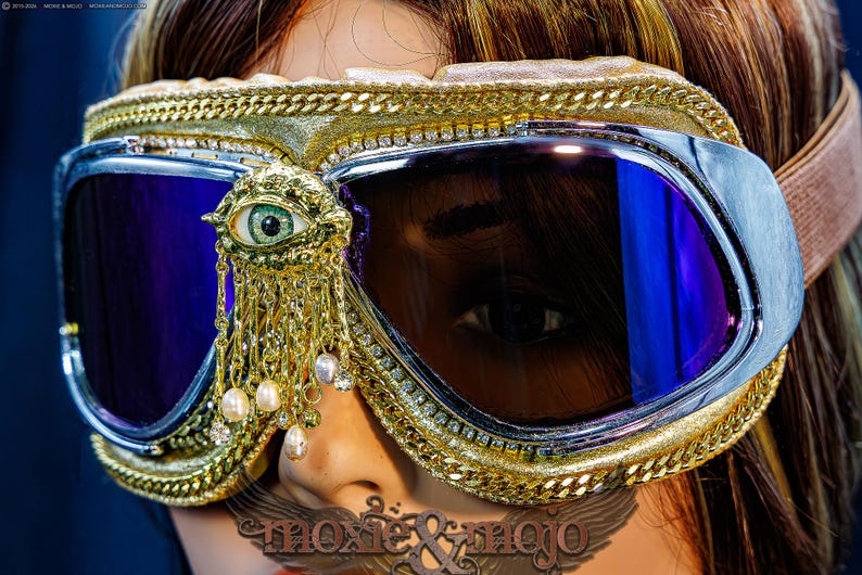 May include: Goggles with dark purple lenses, a gold-toned frame, and a decorative gold eye emblem featuring dangling white pearls and clear crystals. The frame is accented with a gold chain trim and a row of small clear rhinestones.