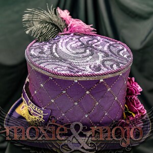 May include: A purple top hat featuring a diamond-patterned side with embedded crystals, a top panel with swirling sequin embroidery, a black and pink feather plume, and a purple masquerade mask accent. The text moxie & mojo is visible.