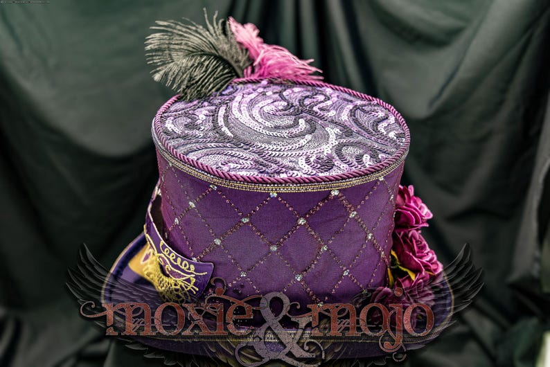 May include: A purple top hat featuring a diamond-patterned side with embedded crystals, a top panel with swirling sequin embroidery, a black and pink feather plume, and a purple masquerade mask accent. The text moxie & mojo is visible.