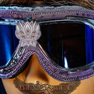 Puede incluir: Gafas protectoras de color morado con lentes reflectantes azul oscuro y un emblema central de flor de loto plateada. El marco est&aacute; decorado con peque&ntilde;os cristales morados y una cadena met&aacute;lica. El texto moxie & mojo es visible abajo.