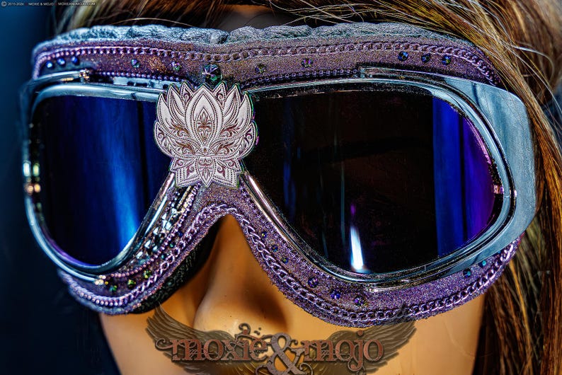 Puede incluir: Gafas protectoras de color morado con lentes reflectantes azul oscuro y un emblema central de flor de loto plateada. El marco est&aacute; decorado con peque&ntilde;os cristales morados y una cadena met&aacute;lica. El texto moxie & mojo es visible abajo.